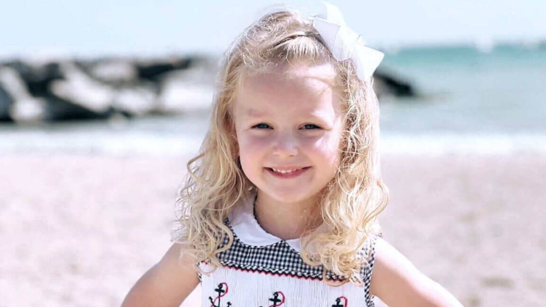 Smiling girl in navy gingham Smocked Anchor Dress from Emily Lacey, enjoying a sunny day at the beach.