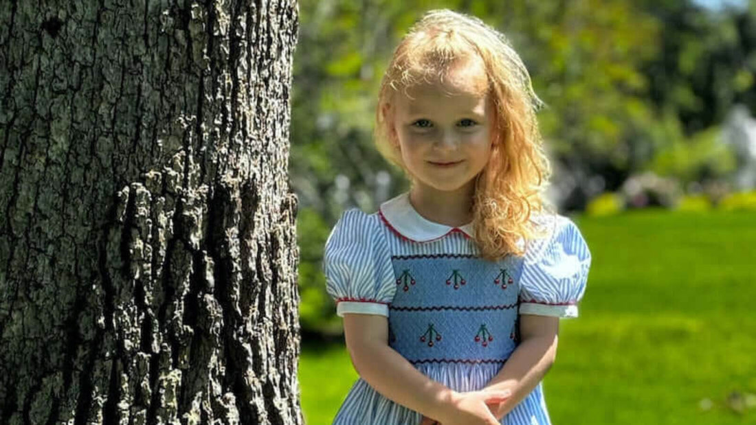 Little girl in classic smocked dress stands by tree, ready for back-to-school in style and comfort.