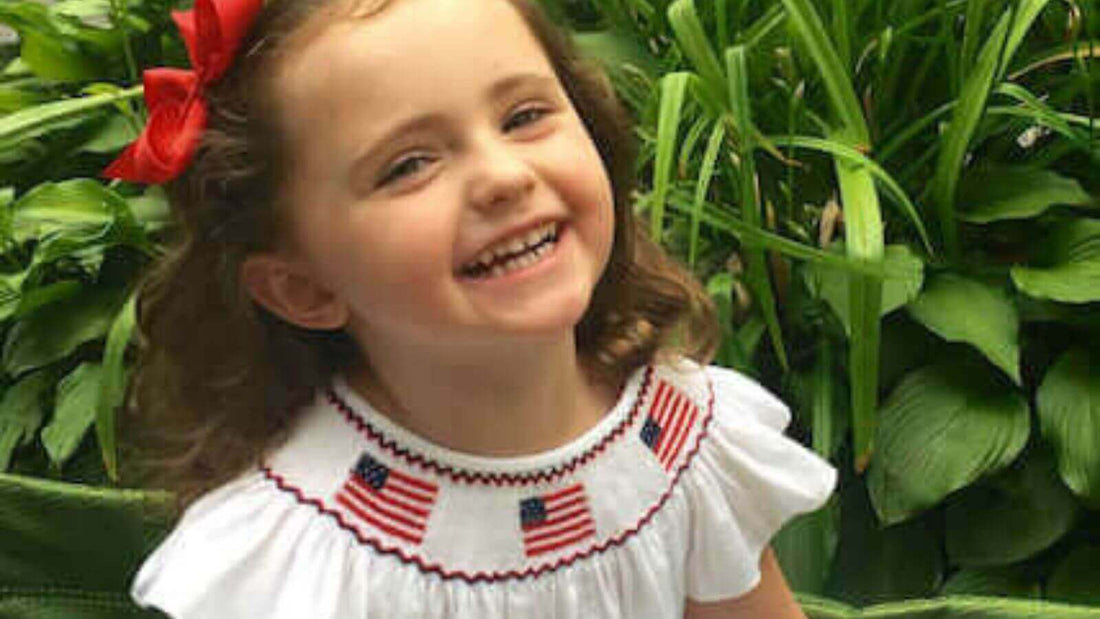 Happy child in a white smocked bodice dress with patriotic flags, wearing a red hair bow, smiling outdoors.