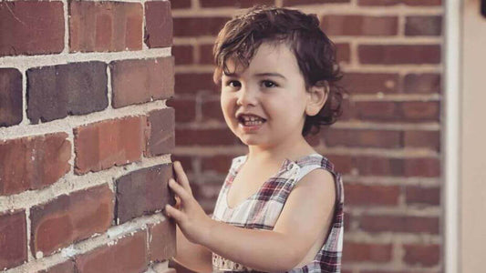 Adorable child in Smocked Tartan John John enjoying timeless comfort and modern style against a rustic brick wall.