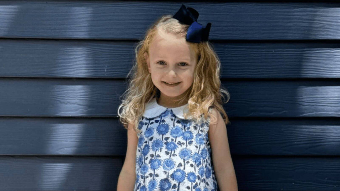 Little girl smiling in stylish Blue Sunflower Dress by Emily Lacey against a blue wooden background