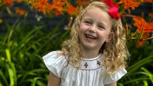 Happy little girl wearing a smocked white cherry bishop dress and red bow, smiling in a garden with vibrant flowers.