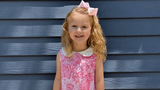 Smiling girl in a pink smocked dress from Emily Lacey standing against a blue wooden wall on picture day.