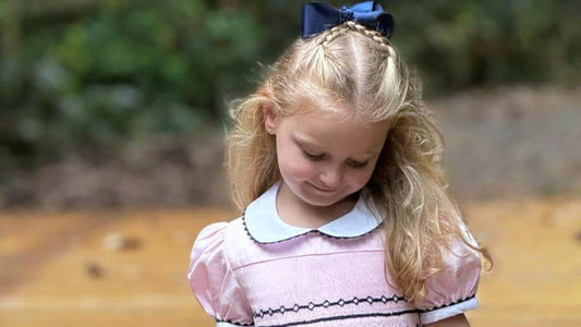 Little girl in a smocked pink and navy geo dress with a navy bow, gazing downwards in an outdoor setting.