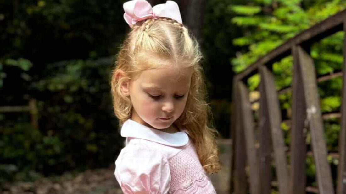 Little girl wearing a smocked pink garland dress with a bow, standing on a bridge in a serene outdoor setting.