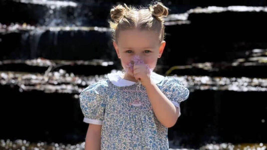 Little girl in a handmade blue smocked dress, holding a flower, and standing in front of a waterfall.