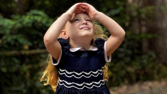 Little girl in a Smocked Navy Garland Dress stands in a forest, gazing upwards with hands on her head, embodying timeless elegance.