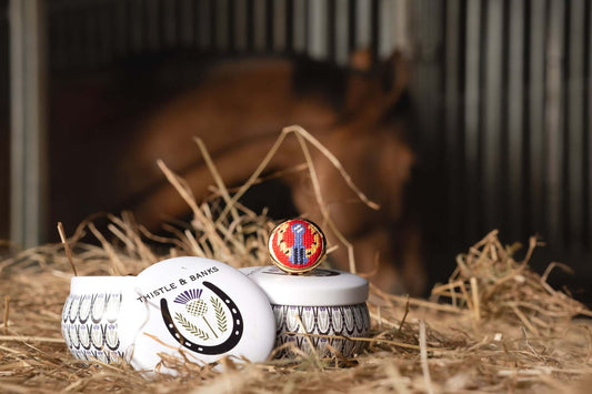 Thistle & Banks needlepoint jewelry resting on hay with a blurred horse in the background, showcasing equestrian elegance.