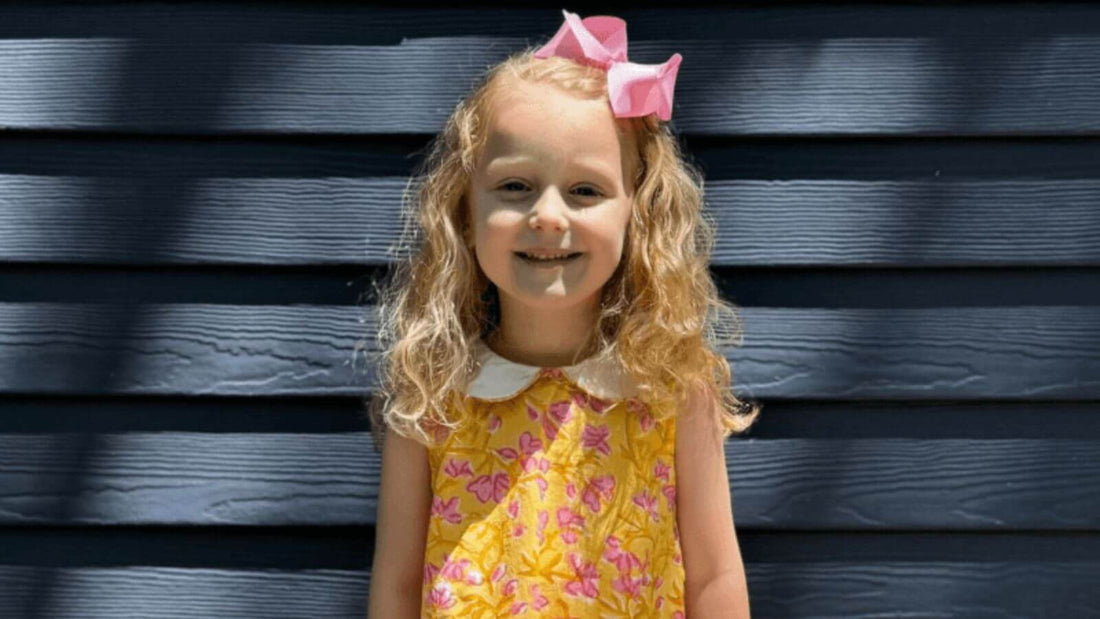 Little girl in a yellow floral dress with a pink bow, smiling against a navy blue wooden background.