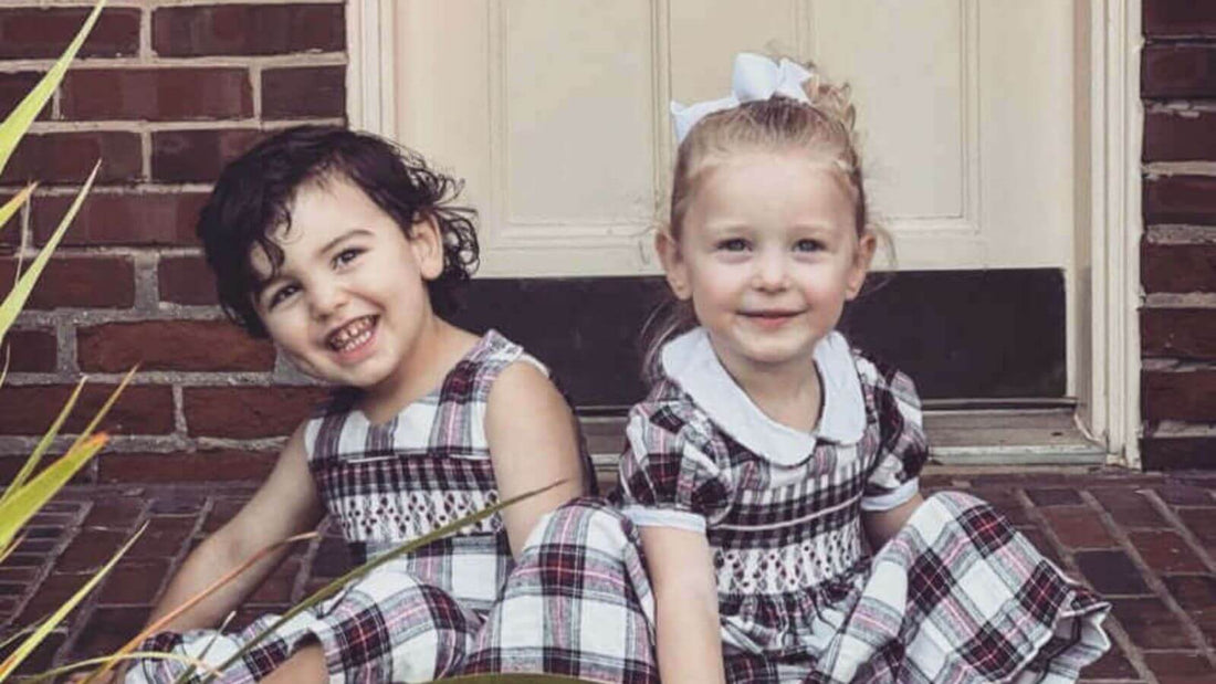 Adorable children wearing Emily Lacey smocked tartan dresses, showcasing timeless fashion with modern style on a brick doorstep.