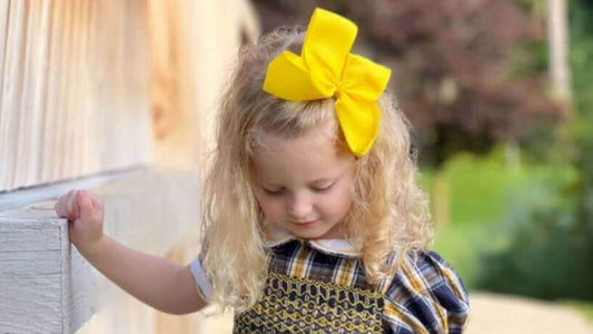 Little girl with curly blonde hair wearing a bright yellow hair bow, showcasing stylish elegance against a serene outdoor backdrop.