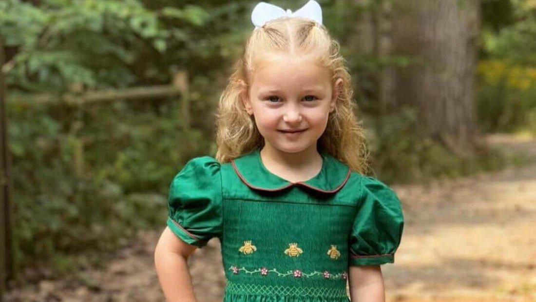 Young girl wearing an elegant green Smocked Royal Bee Dress with intricate hand-smocked bee designs and a white bow in her hair