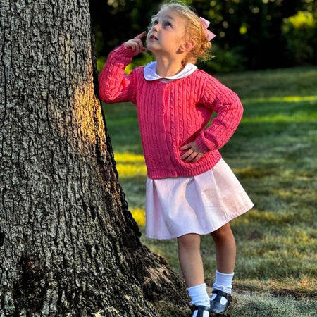 Little girl in a pink cable knit cardigan and white skirt poses by a tree, embodying timeless children's fashion charm.