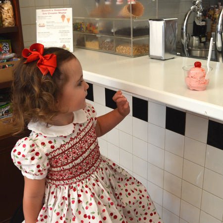 Little girl in cherry print smocked dress at ice cream shop, promoting a 30% discount by ordering by 2/28.