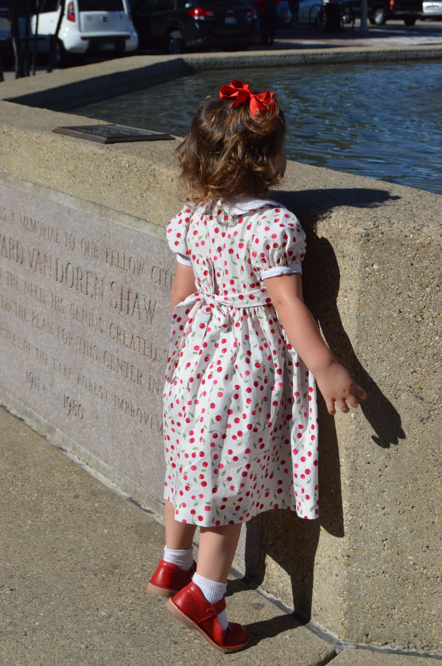 Little girl in a smocked cherry dress by a fountain, enjoying a sunny day.