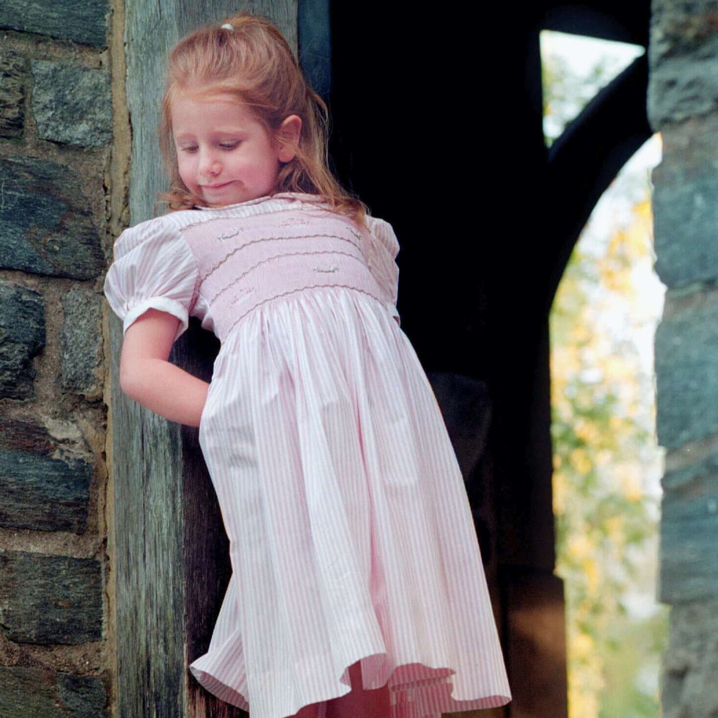 Girl in a pink striped hand-smocked dress with lovebirds, posed against a stone wall, showcasing comfort and charm.