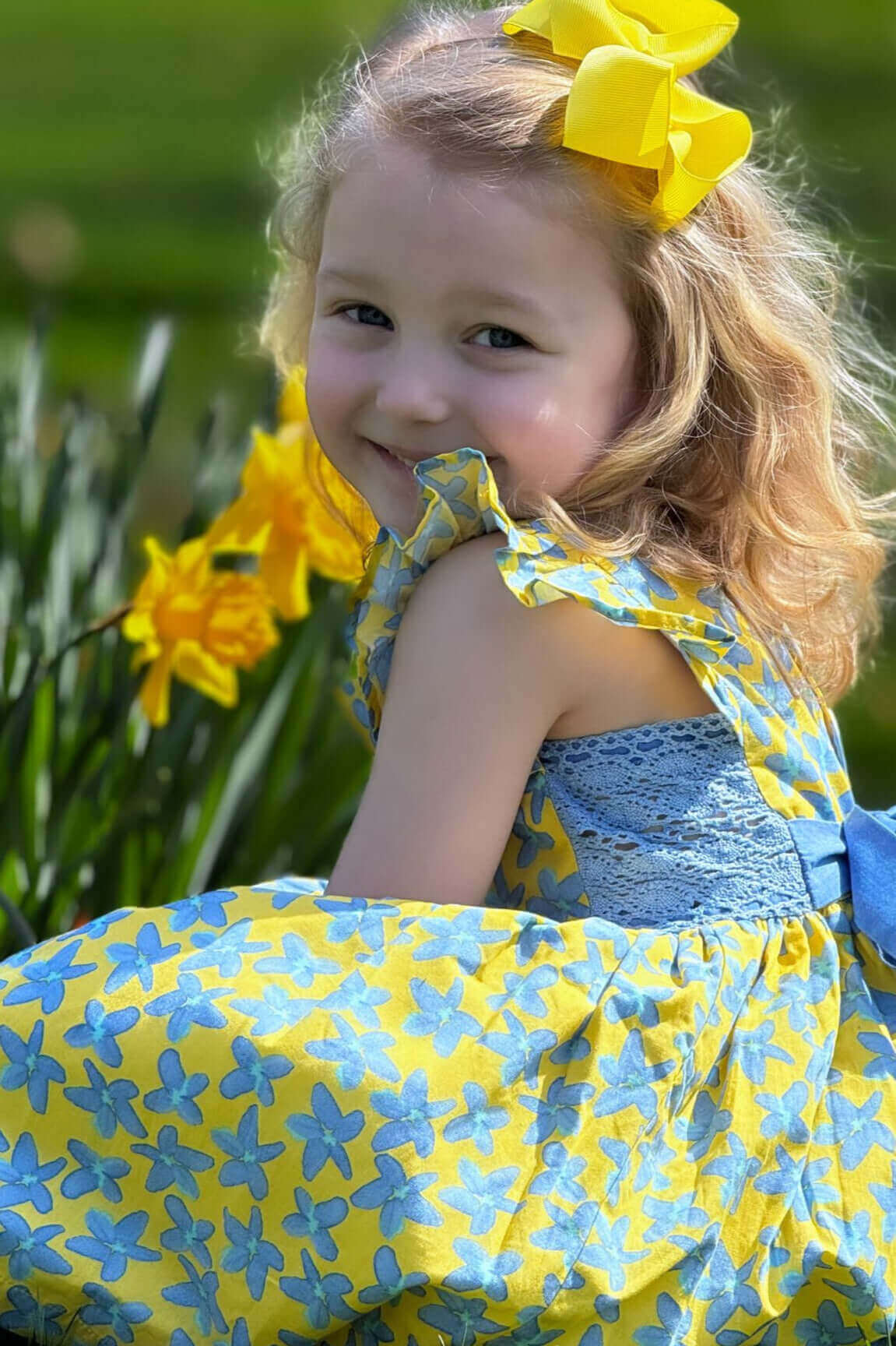 Little girl in a Forget Me Not Dress smiles among spring flowers, wearing a yellow and blue floral print dress with a cornflower belt.