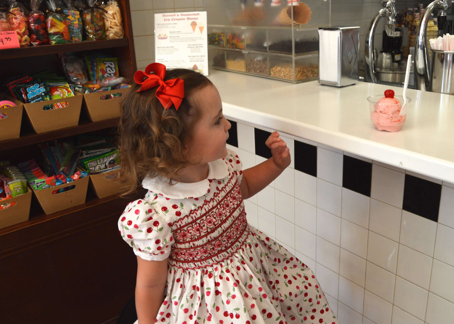 Little girl in a smocked cherry dress enjoying ice cream in a charming candy shop.