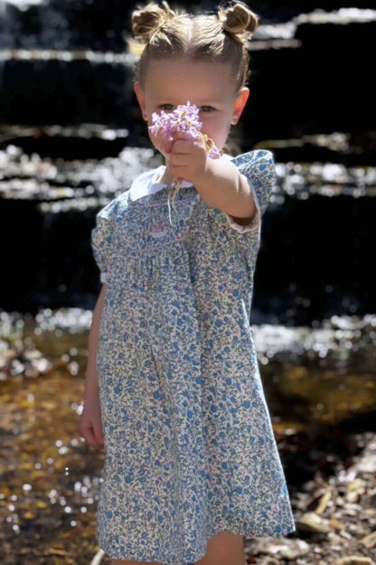 Child in Smocked Flower Basket Dress holding flowers by a stream, showcasing intricate blue floral design and enchanting embroidery.