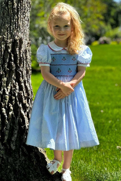 Little girl wearing a Smocked Striped Cherry Dress with cherry embroidery and ric rac trim, standing by a tree on a sunny day.