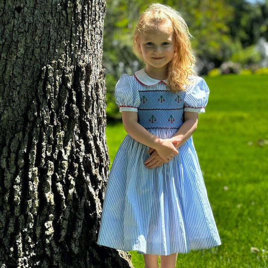 Little girl in a blue dress standing by a tree on a sunny day.