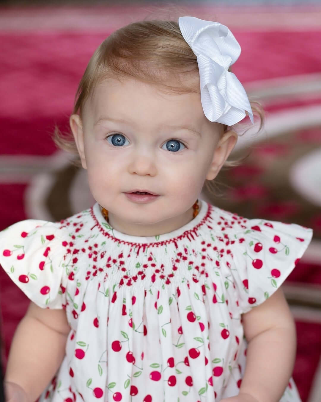 Sweet baby girl wearing a smocked cherry dress with a white bow, showcasing the adorable cherry print and delicate neckline.