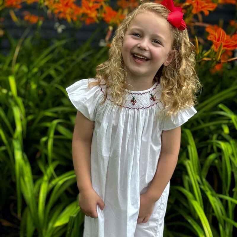 Smocked cherry bishop dress on a smiling girl with curly hair standing in a garden, showcasing delicate cherry embroidery.