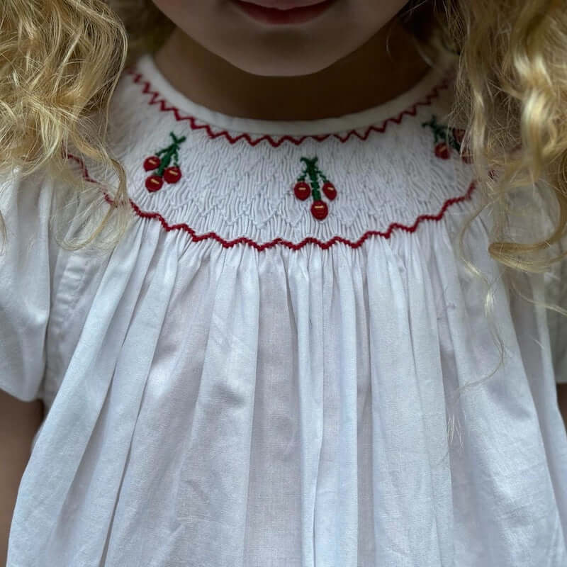 Close-up of hand-smocked cherry bishop dress featuring red cherry embroidery on a white cotton fabric, perfect for toddlers.