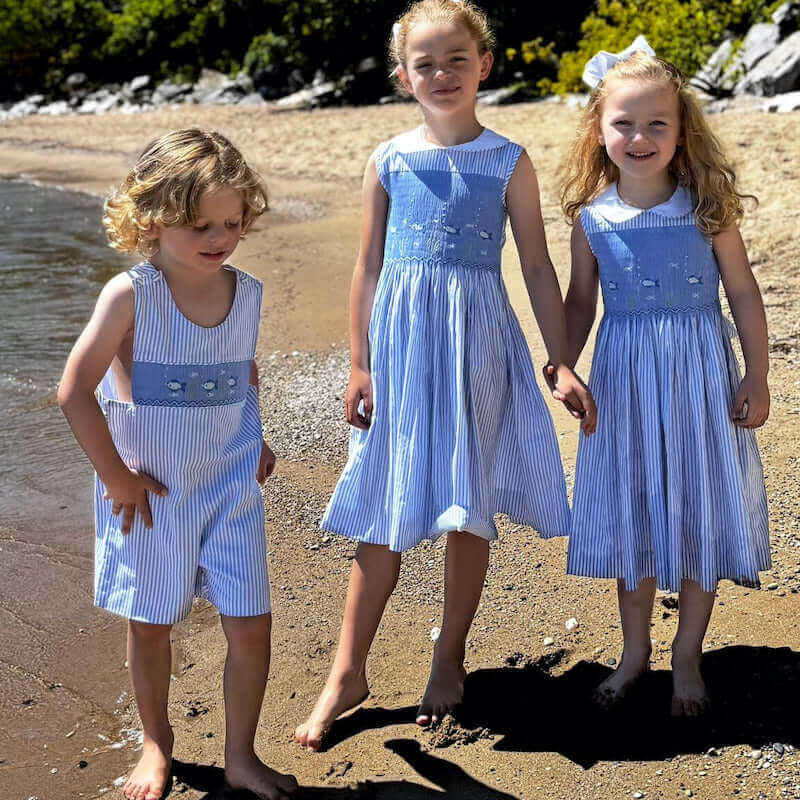 Children on the beach wearing Smocked Fish John John outfits in classic blue stripes, showcasing maritime charm.