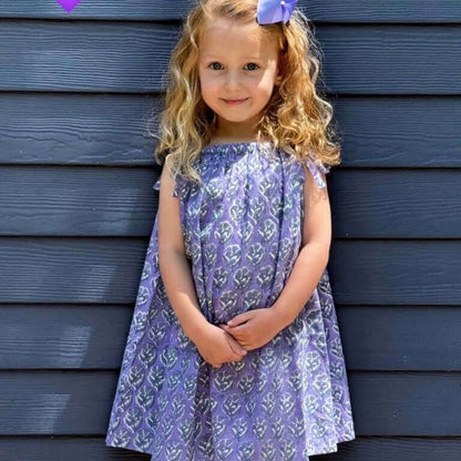 Little girl wearing a cute purple floral sun dress, perfect for school, posing with charming smile in front of a blue wall.