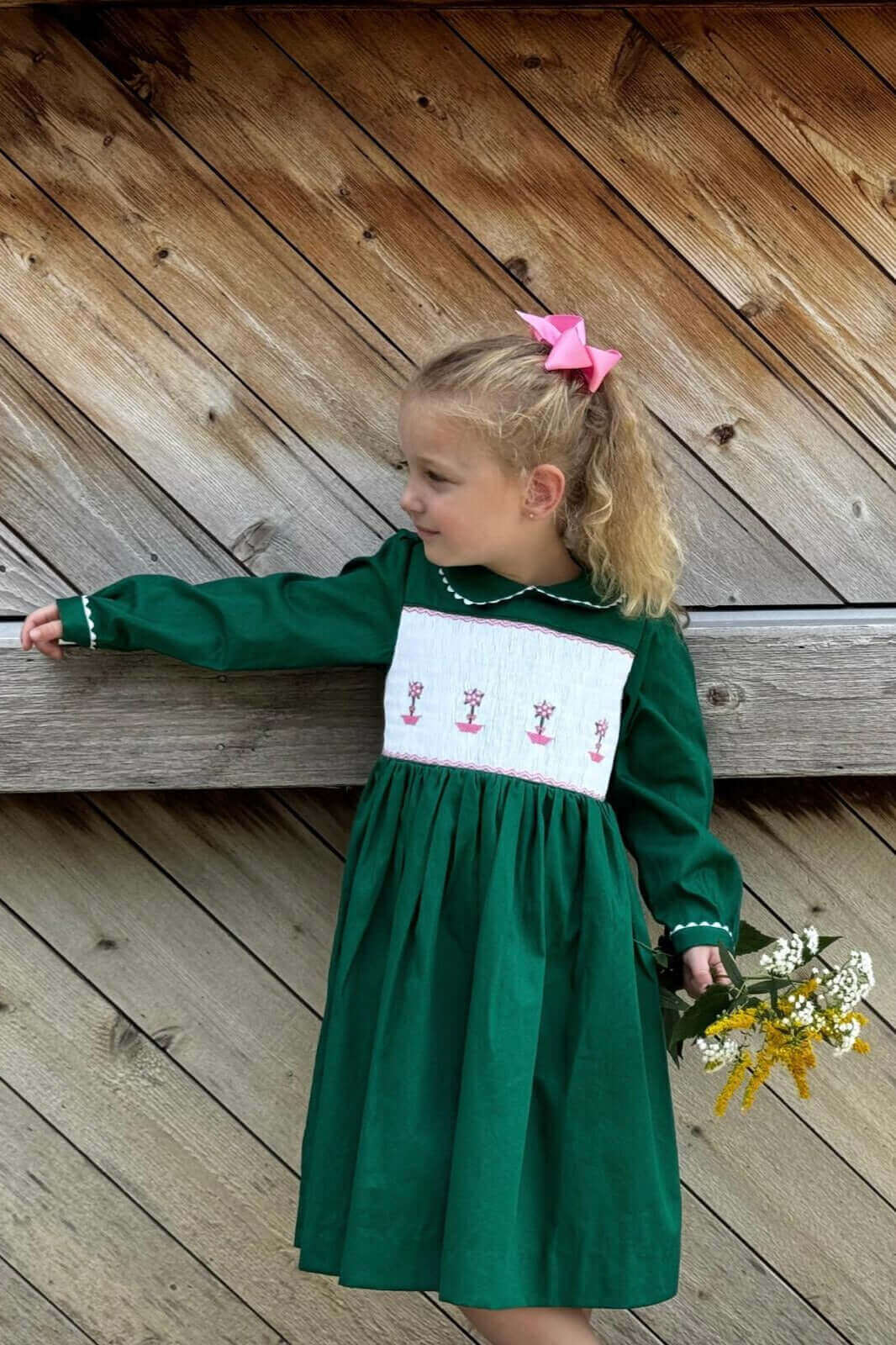 Little girl in a smocked green topiary dress with pink bow, holding flowers. Perfect for school photos, birthdays, holidays and daily wear.