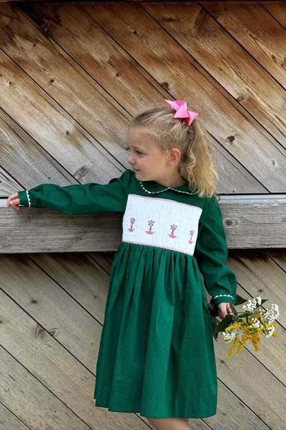 Little girl in a smocked green topiary dress with pink bow, holding flowers. Perfect for school photos, birthdays, holidays and daily wear.