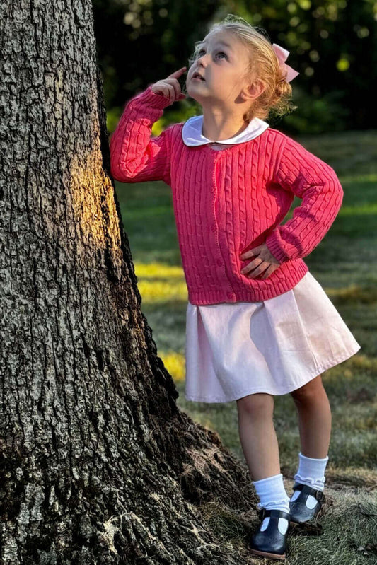 A little girl poses thoughtfully by a tree, wearing a hot pink cable knit cardigan and white dress.