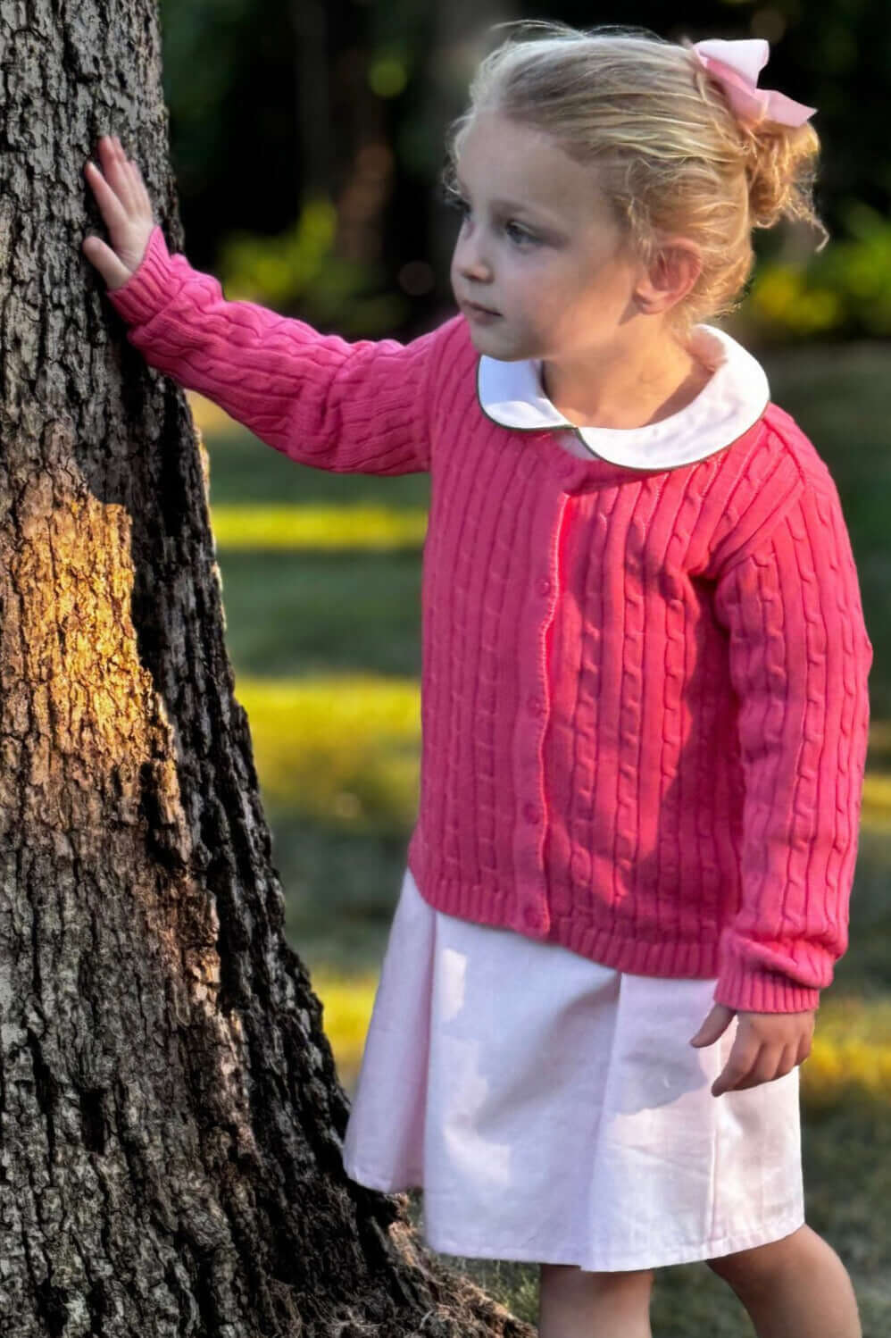 Young girl in a pink cable-knit sweater and white dress stands by a tree, enjoying a sunny day outdoors.