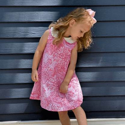 Young girl in pink watercolor A-line dress with Peter Pan collar, button back, and attached belt, standing in front of blue wall.