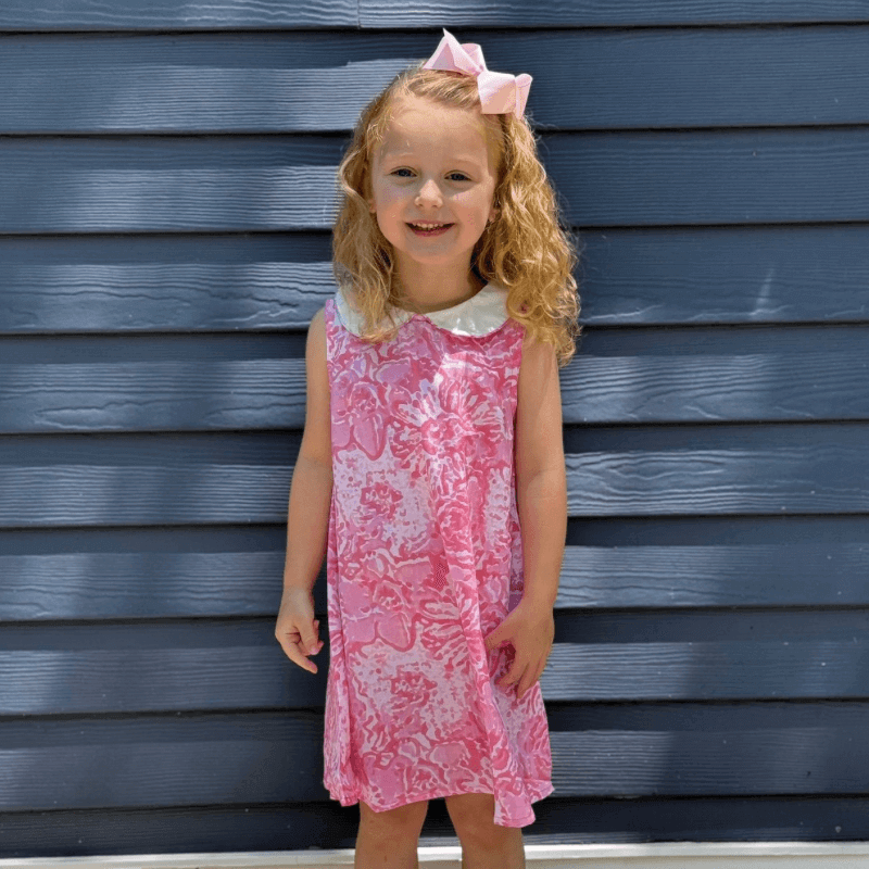 Little girl in a pink watercolor dress with Peter Pan collar and A-line cut, smiling in front of a blue wooden wall.