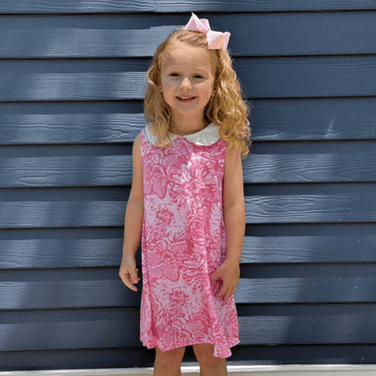 Little girl in a pink watercolor dress with Peter Pan collar and A-line cut, smiling in front of a blue wooden wall.