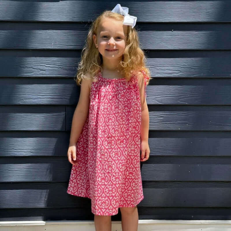 Adorable girl in a pink floral sun dress smiles in front of a dark blue wall.