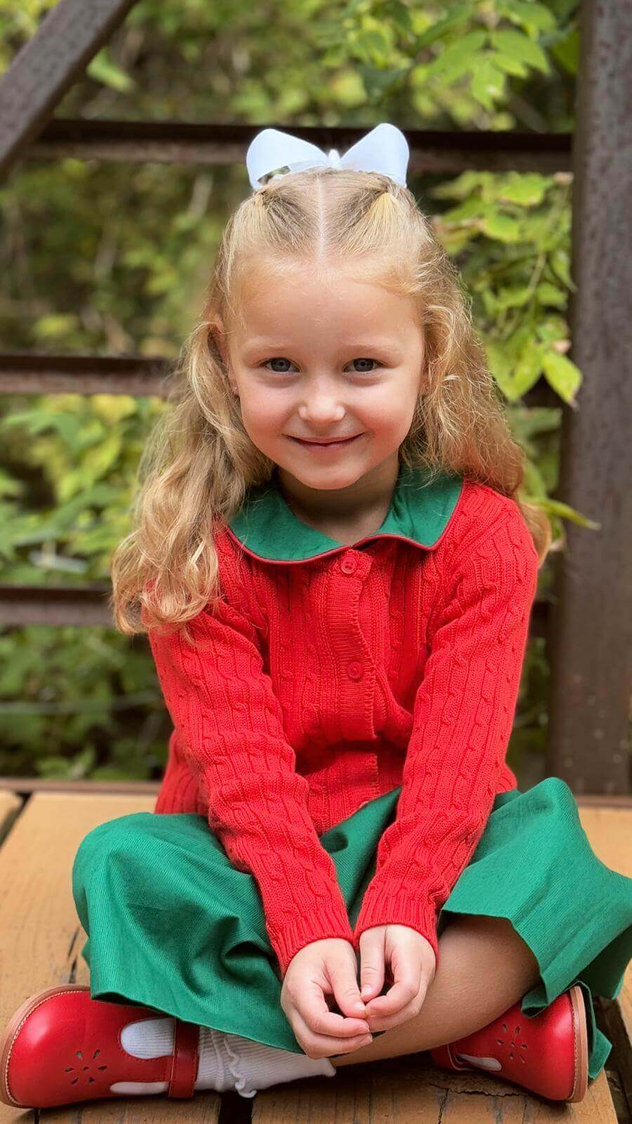 Little girl smiling in a red cable knit cardigan, green dress, and white bow headband sitting outdoors.