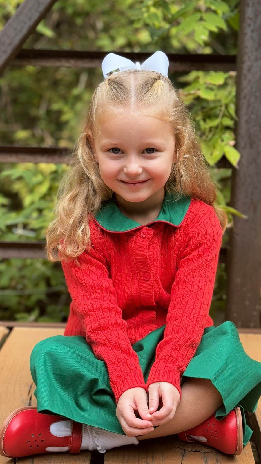 Adorable girl in a red sweater and green skirt, smiling happily with a white bow in her hair, sitting outdoors.
