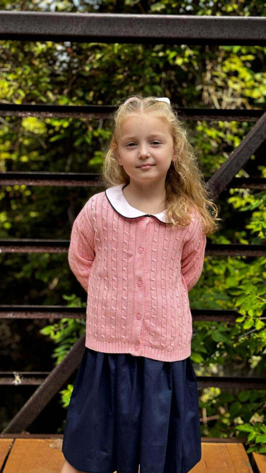 Little girl in pink organic cotton cable knit cardigan and navy skirt, standing outdoors in front of greenery.