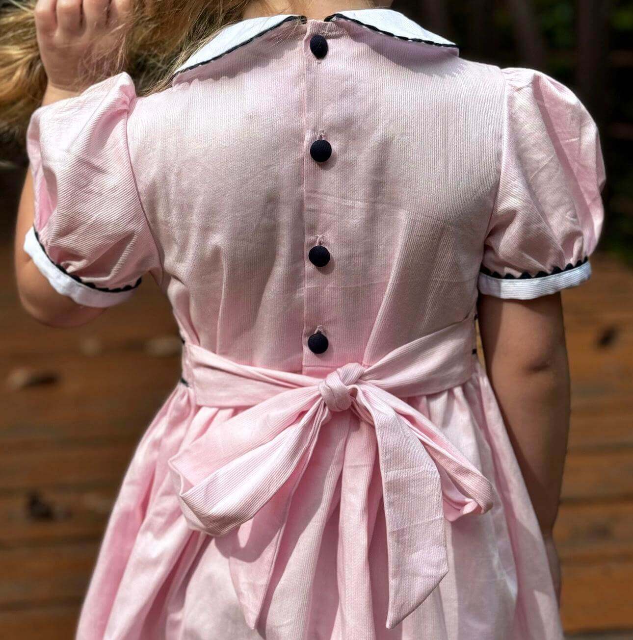 Rear view of a girl wearing a hand-smocked pink and navy geo dress with covered buttons and ric rac trim.