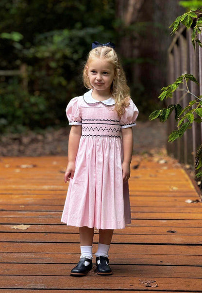 A young girl wearing a Smocked Pink and Navy Geo Dress stands on a wooden path in a forest, showcasing elegant handcrafted details.