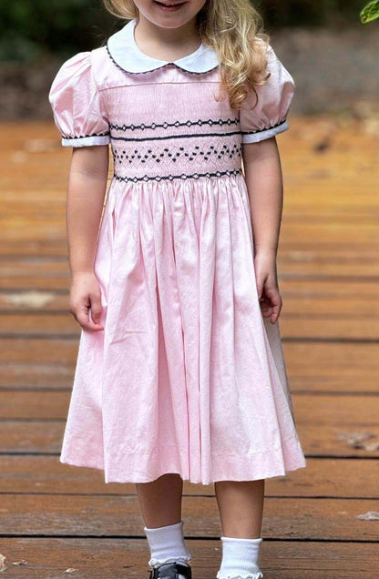 Toddler in a smocked pink and navy geo dress with puff sleeves and ric rac trim standing on a wooden path.