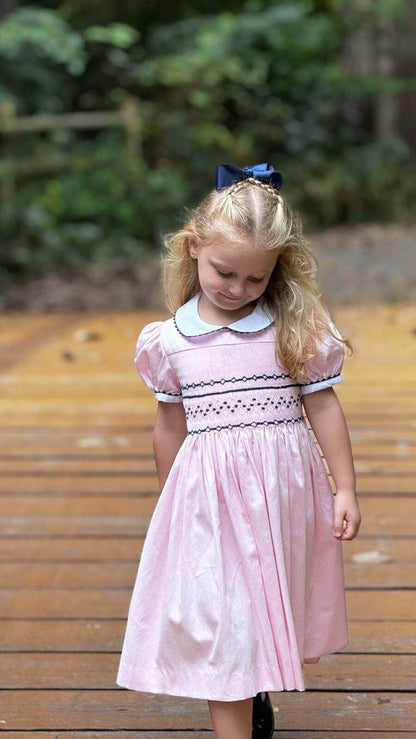 Little girl wearing a hand-smocked pink and navy geo dress, perfect for school photos, birthdays, holidays, or daily wear, with sophisticated details.