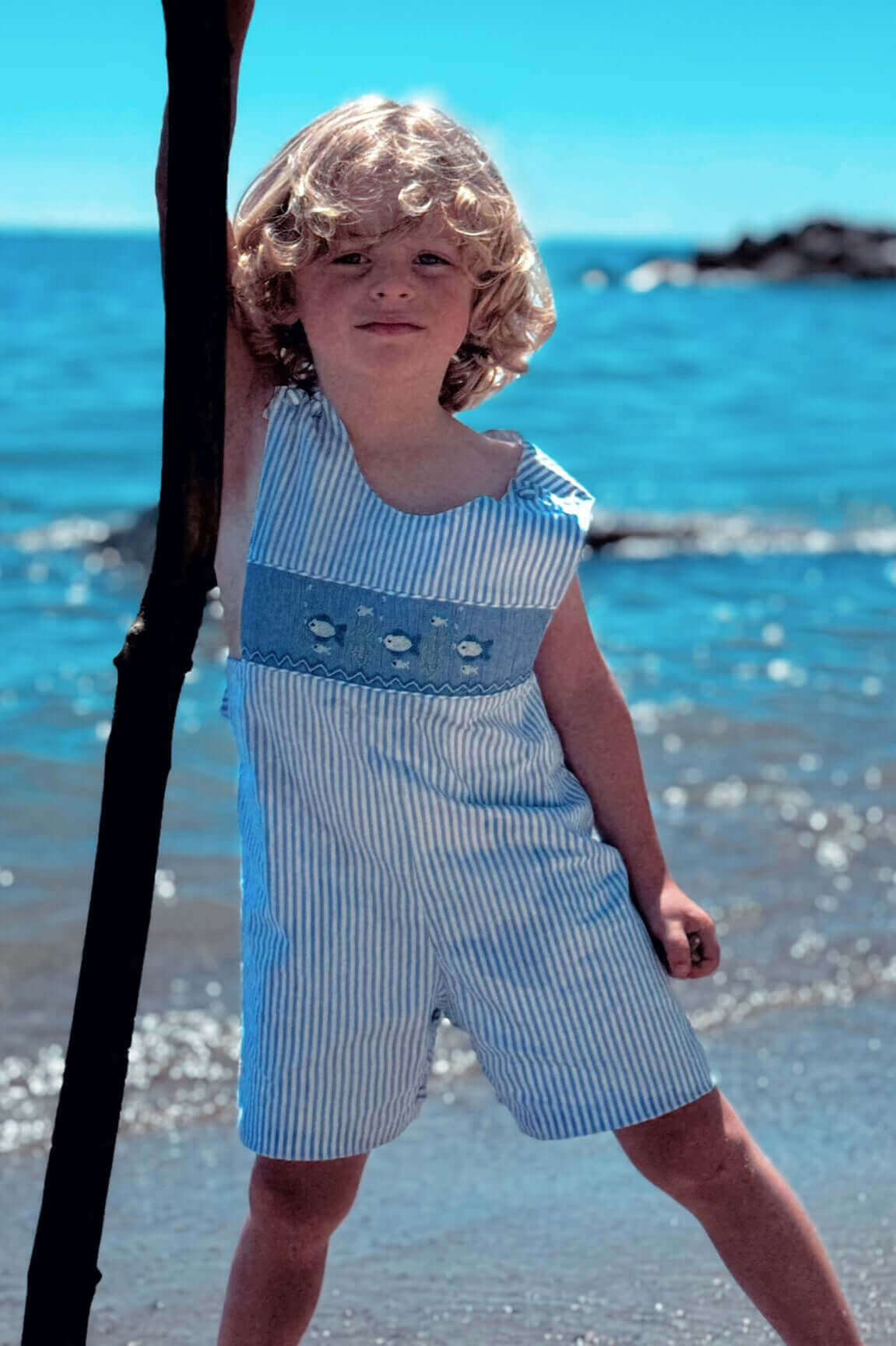 Smocked Fish John John in classic french blue stripe worn by a child at the beach, enjoying summer fun.