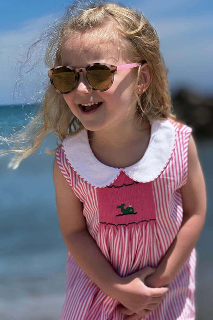 Little girl in a pink and white Smocked Whale Dress, enjoying a sunny day at the beach with stylish sunglasses.