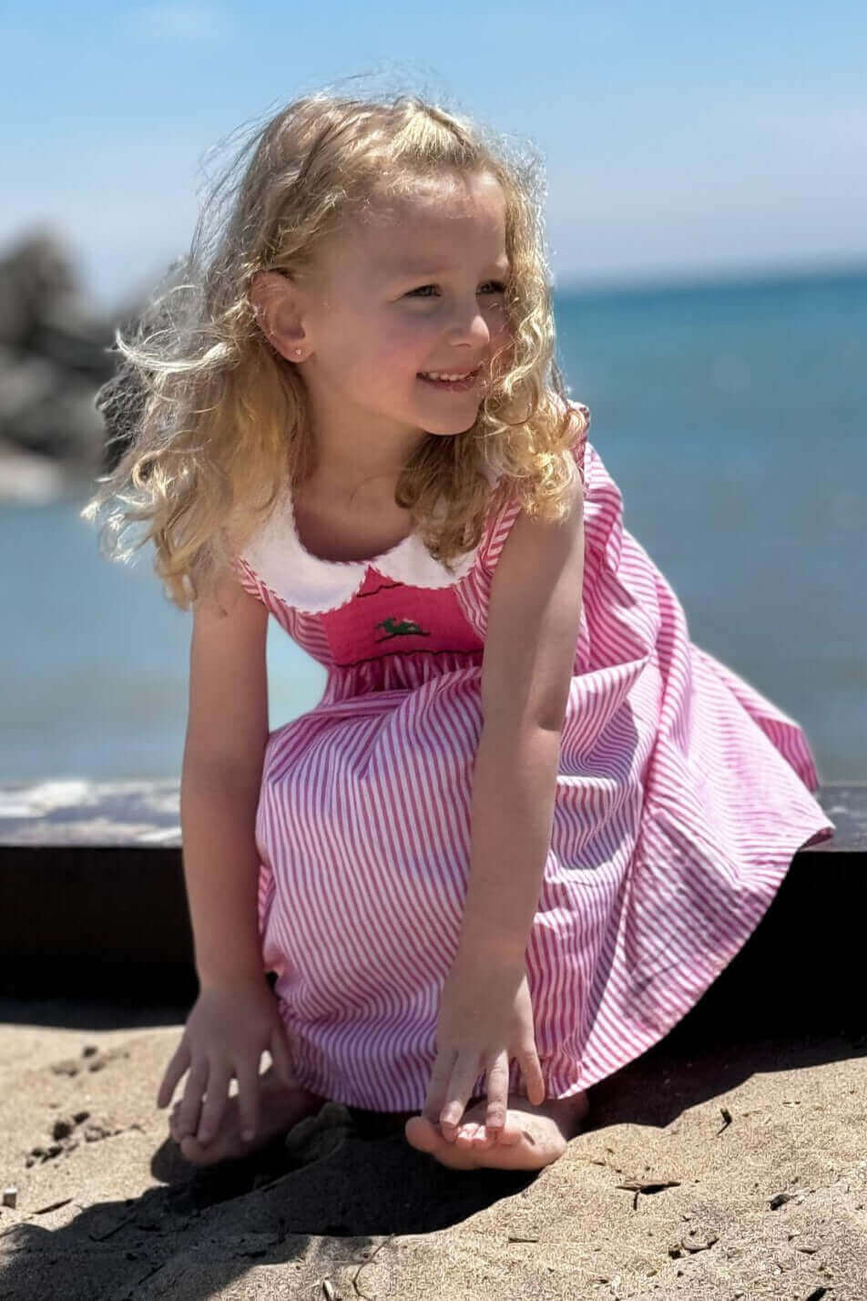 Little girl in a pink and white striped smocked whale dress on the beach, enjoying a sunny day by the sea.