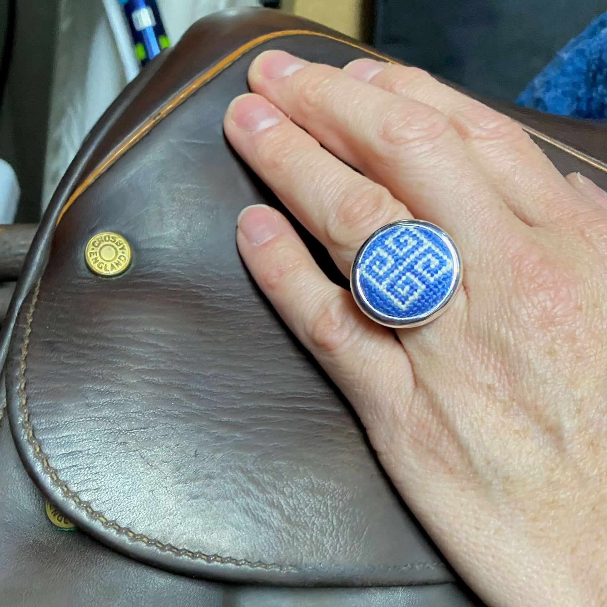 Adjustable periwinkle blue Greek key needlepoint ring worn on hand, resting on a brown leather bag.