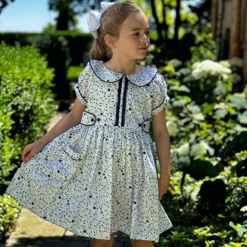 Little girl wearing a Jane Ruffle Dress in blue floral print with Peter Pan collar and ruffled placard, standing in a blooming garden.